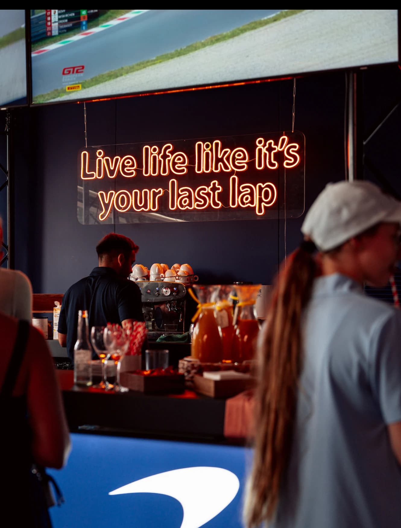 Guests at the hospitality bar under a neon sign reading Live life like it's your last lap