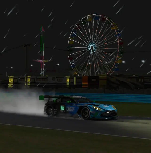 A blue and green GT car throwing up spray under floodlights in night-time rain, a lit big wheel behind the barriers