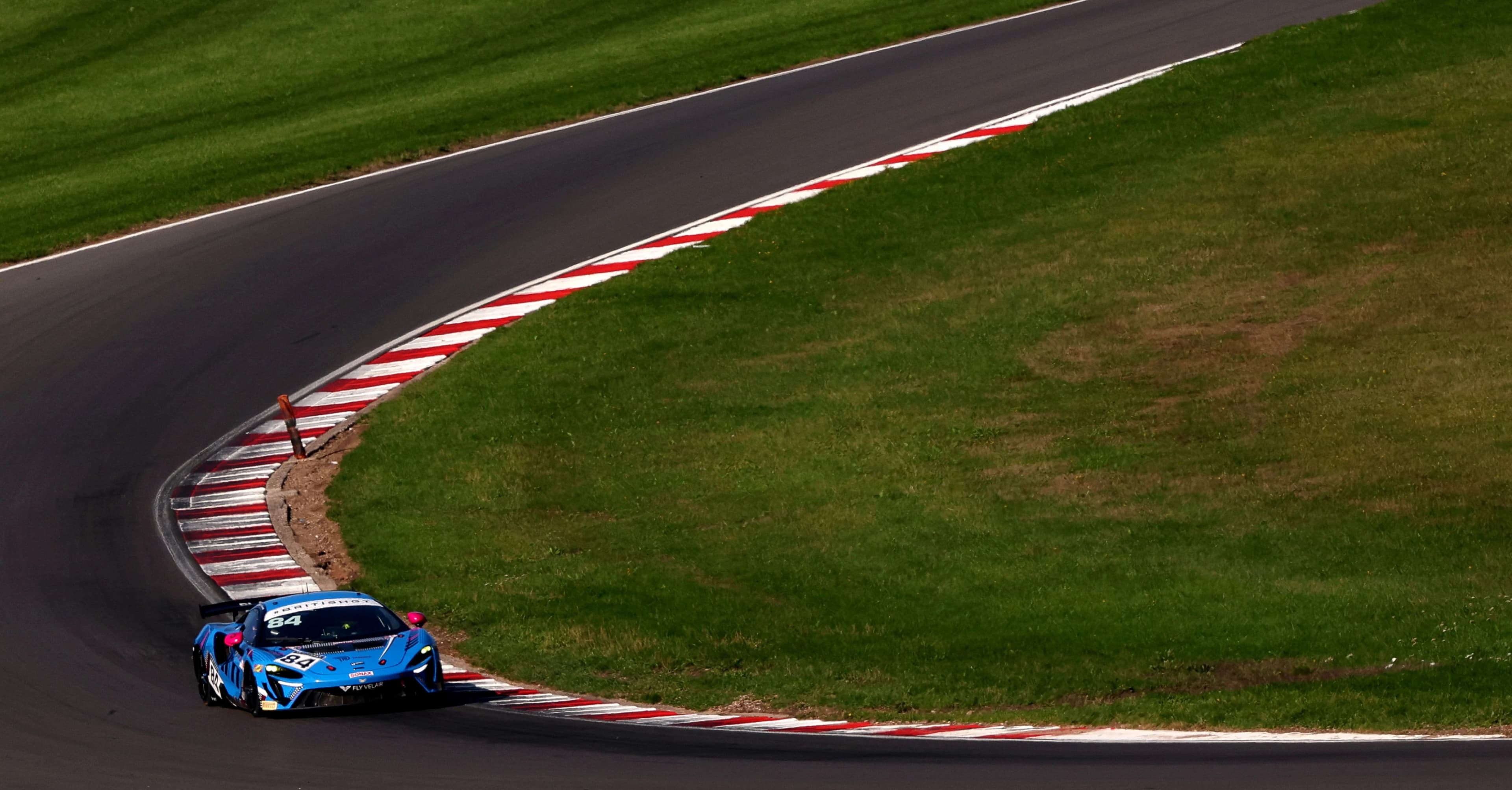 The blue number 84 McLaren Artura GT4 turning in over the red and white kerbs of a sweeping corner
